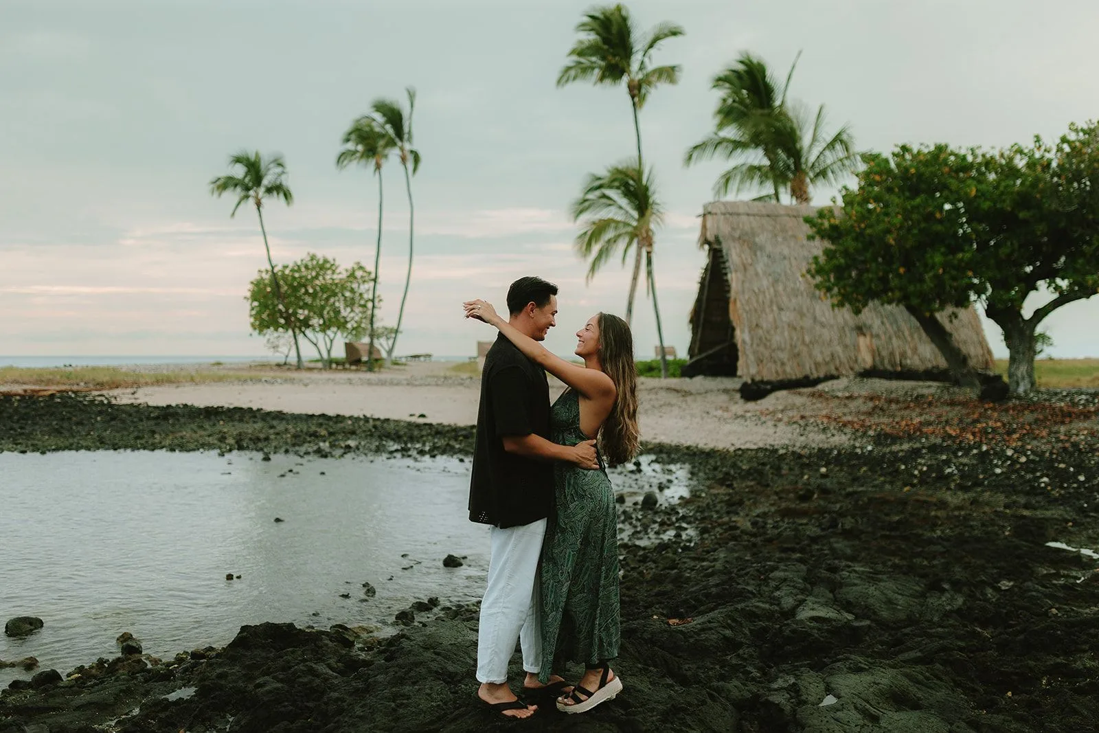 A couple holding each other on a black-lava shoreline beneath swaying palms